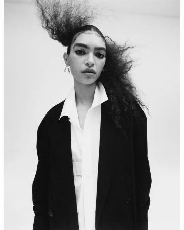 Black and white portrait of a person with gravity-defying hair, wearing a white collared shirt and dark blazer in a light studio.