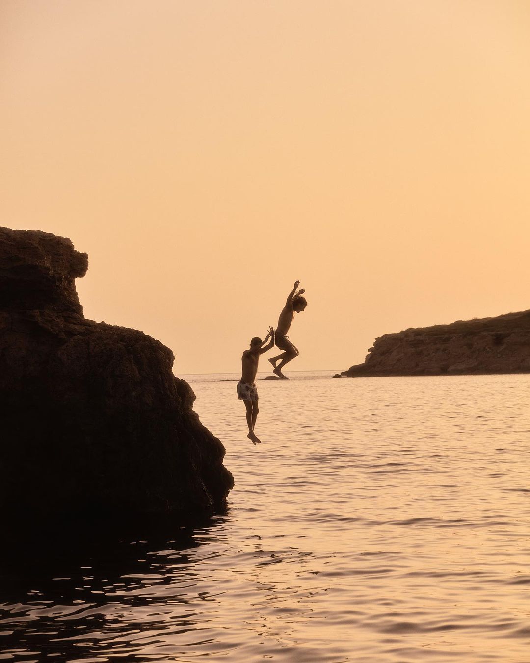 Two young men on a rocky coast at dusk, one pushing the other into the water. Silhouette against orange sky and calm ocean.