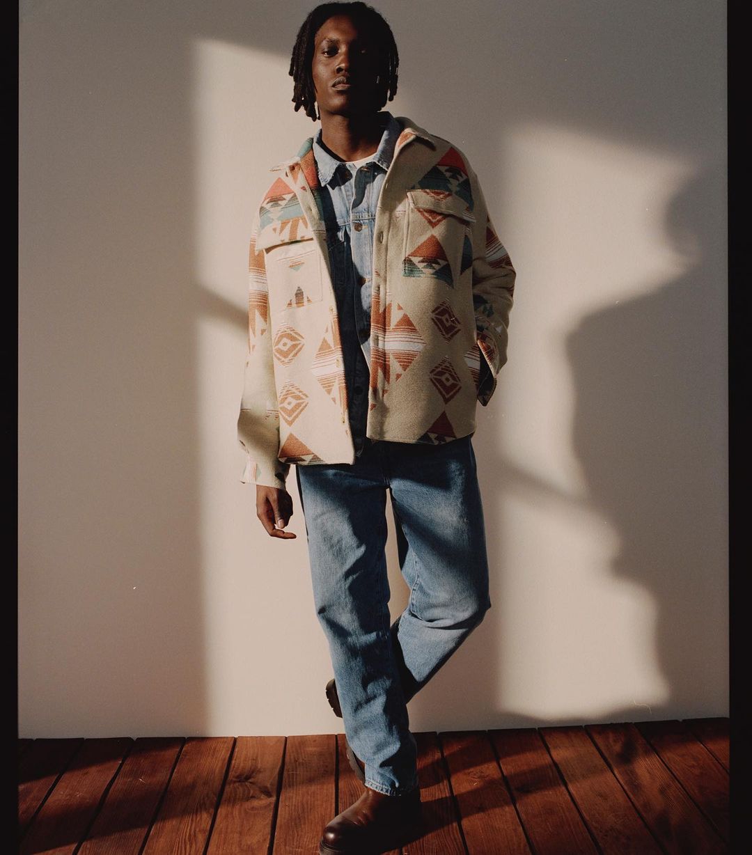 A stylish adult male with dreadlocks, wearing a patterned overshirt, blue denim shirt, jeans, and shoes stands in a photo studio.