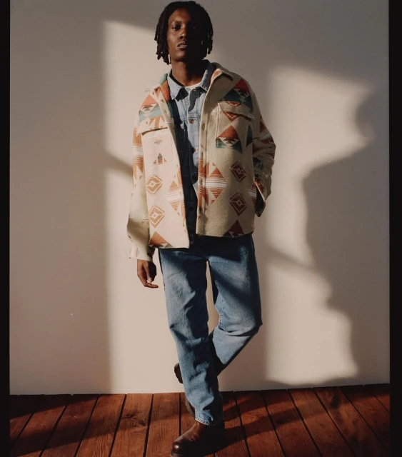 A stylish adult male with dreadlocks, wearing a patterned overshirt, blue denim shirt, jeans, and shoes stands in a photo studio.