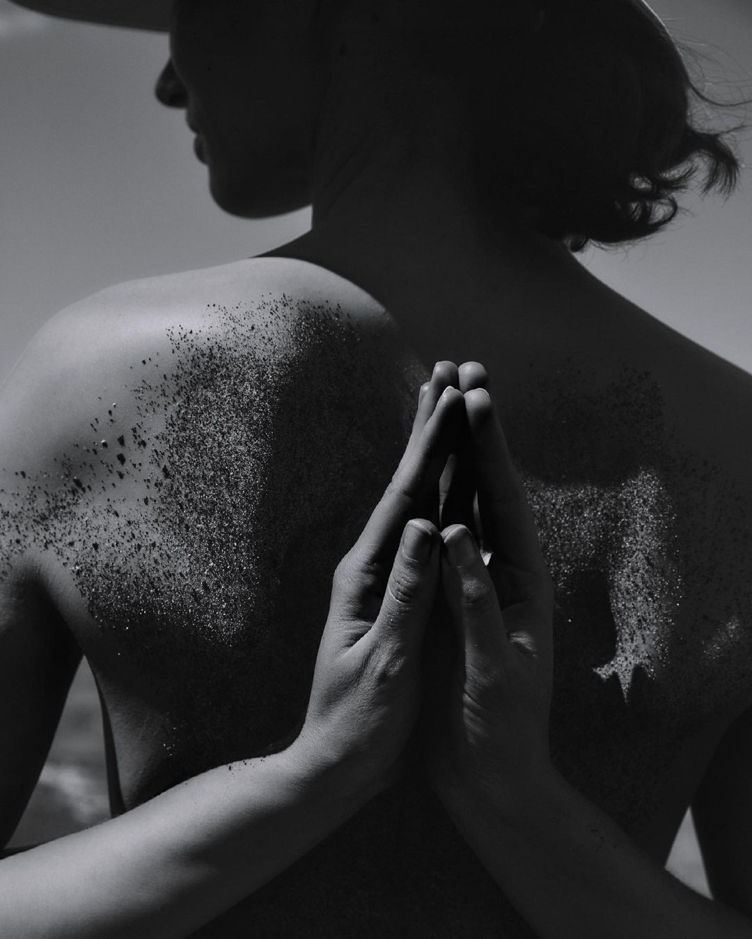 Monochrome portrait of a person with dark hair, back to the viewer, glitter on their shoulders, and hands clasped in a prayer position.
