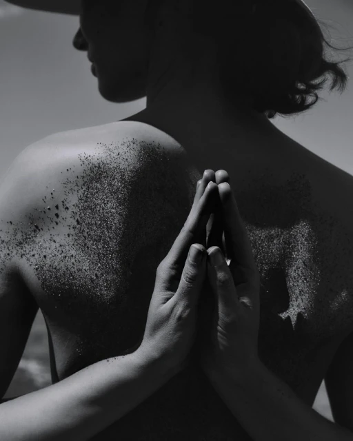 Monochrome portrait of a person with dark hair, back to the viewer, glitter on their shoulders, and hands clasped in a prayer position.