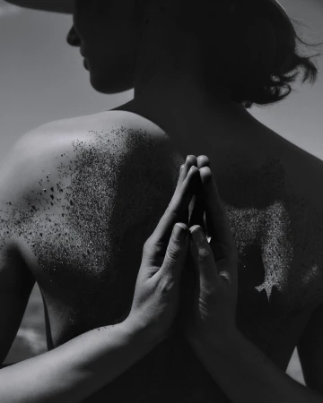 Monochrome portrait of a person with dark hair, back to the viewer, glitter on their shoulders, and hands clasped in a prayer position.