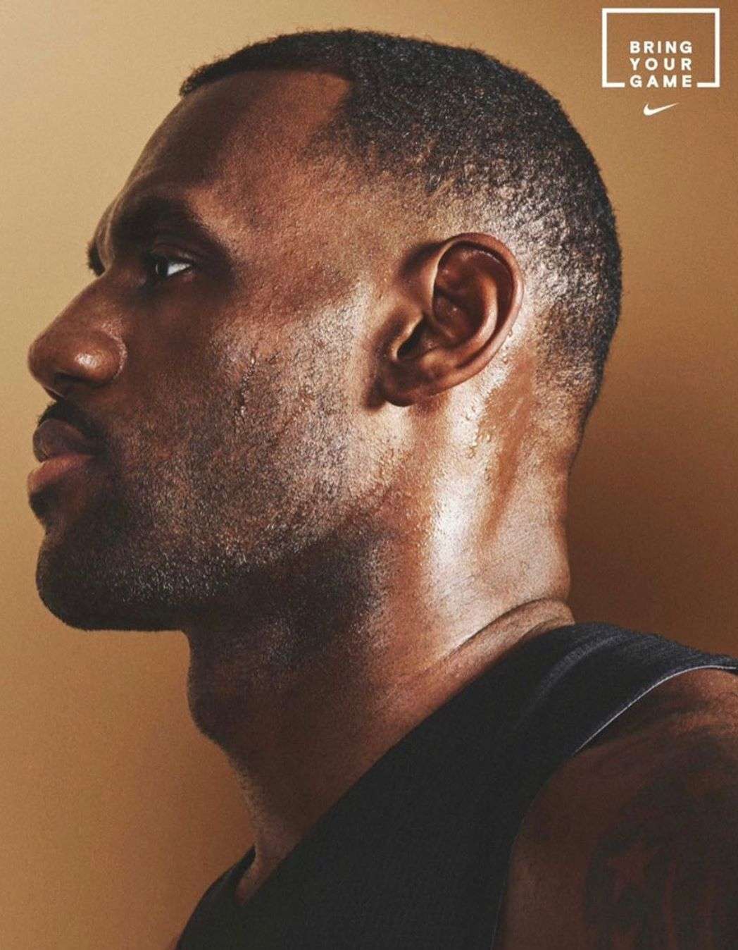 Profile of a sweaty Black adult male with a shaved head and goatee, wearing a black tank top against a neutral background. 