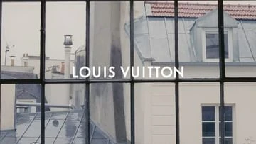 Louis Vuitton logo on a window overlooking a Parisian cityscape with rooftops and dormer windows on a cloudy day. 
