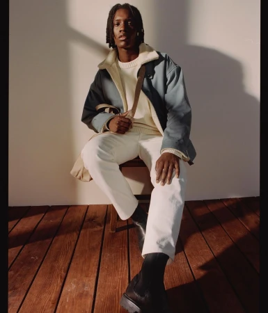 Black man with dreadlocks seated on wooden chair wearing blue jacket, white sweater, and white trousers against wall