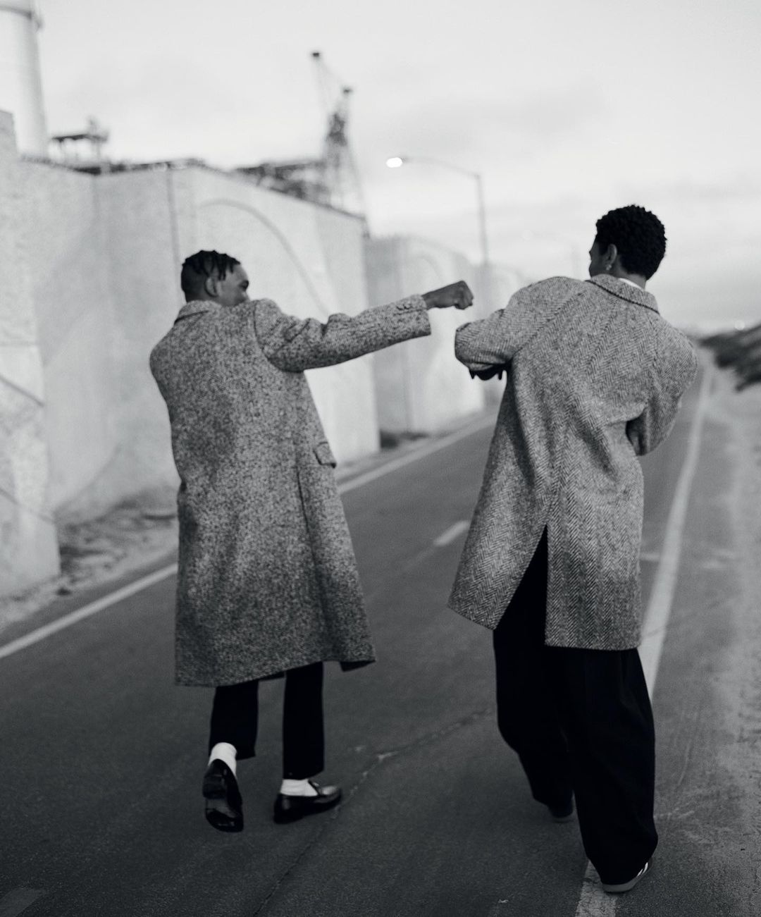 Two adult men in coats walk away on an asphalt road, industrial buildings and cloudy sky in background, black and white photo.