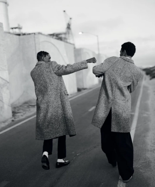 Two adult men in coats walk away on an asphalt road, industrial buildings and cloudy sky in background, black and white photo.