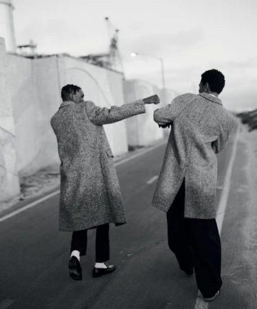 Two adult men in coats walk away on an asphalt road, industrial buildings and cloudy sky in background, black and white photo.