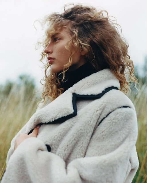 Portrait of a young fair-skinned person with curly blonde hair wearing a light-colored sheepskin coat in a field of tall grass.