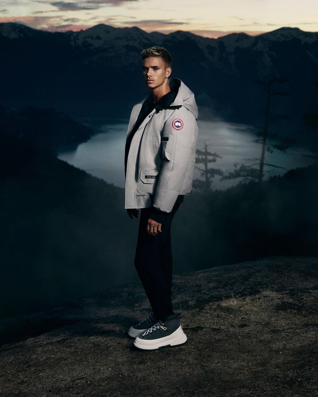 Full-length shot of a male fashion model posing on a rocky hilltop, wearing a grey Canada Goose parka jacket against a mountain backdrop.