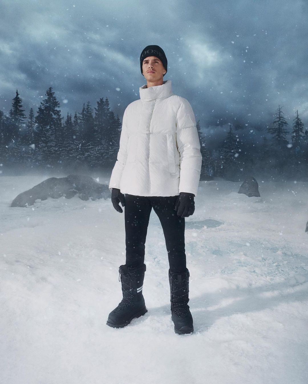 A man stands in a snowy landscape wearing a white puffer jacket, black pants, gloves and beanie, and winter boots, outdoors.