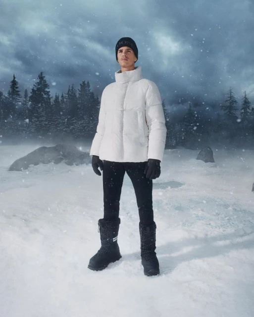 A man stands in a snowy landscape wearing a white puffer jacket, black pants, gloves and beanie, and winter boots, outdoors.