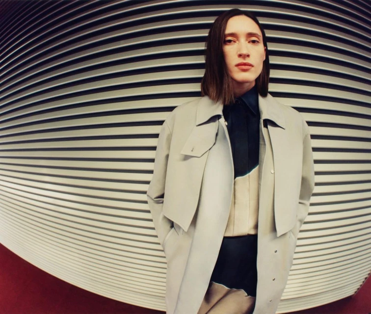 Adult poses in a light tan coat against a black and white striped background with red floor in a fashion editorial photo.