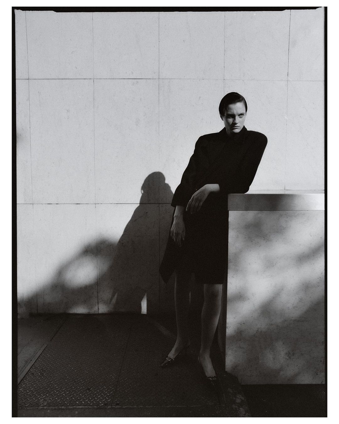 Model in dark clothing leaning against a tiled architectural wall with strong shadows by photographer Misha Taylor.
