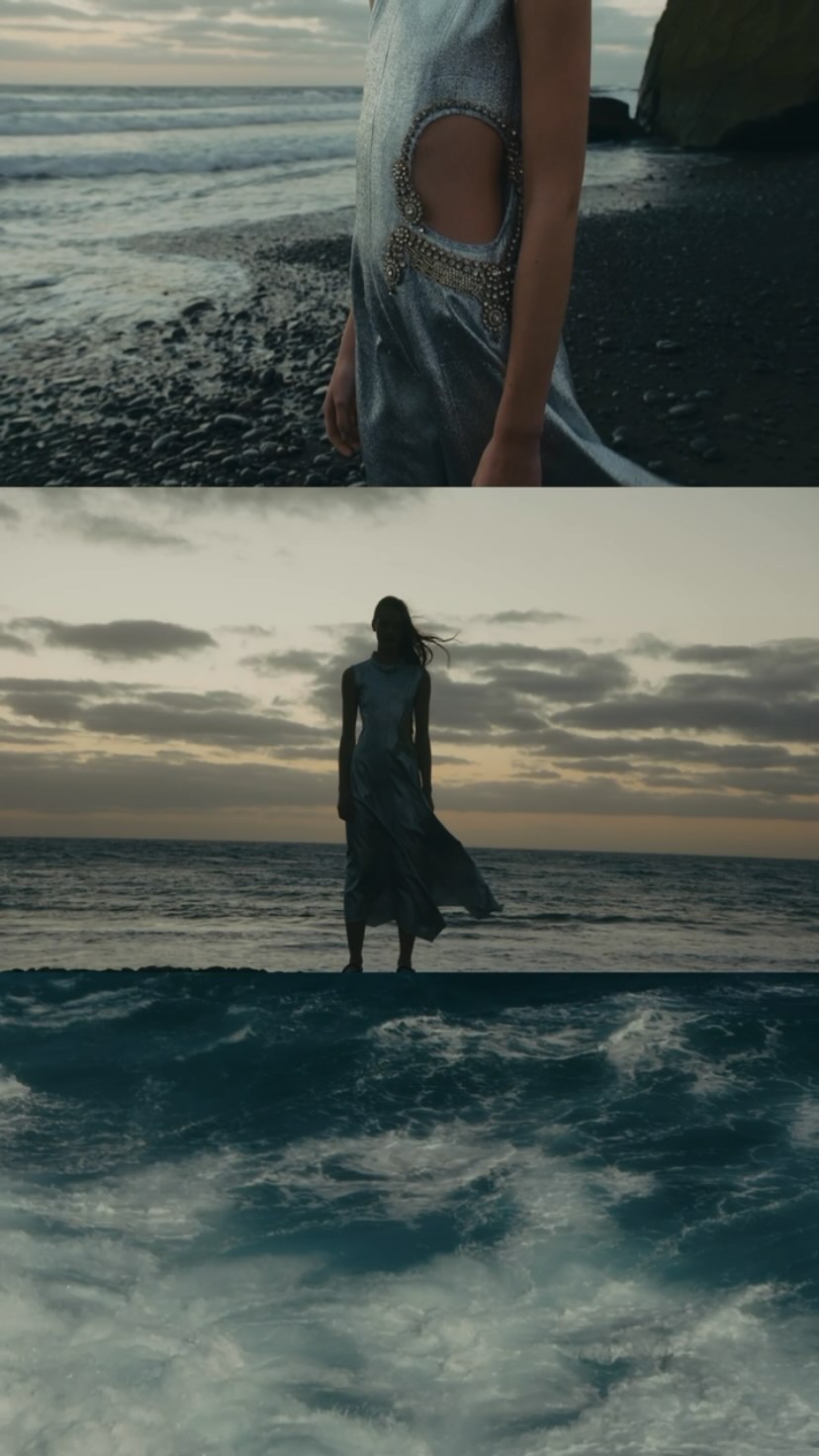 Model in a silver cutout dress standing on a dark pebble beach by the ocean at dusk, photographed by Misha Taylor.