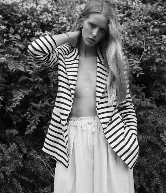 Black and white portrait of a woman with long blonde hair wearing a striped blazer and standing against a leafy background.