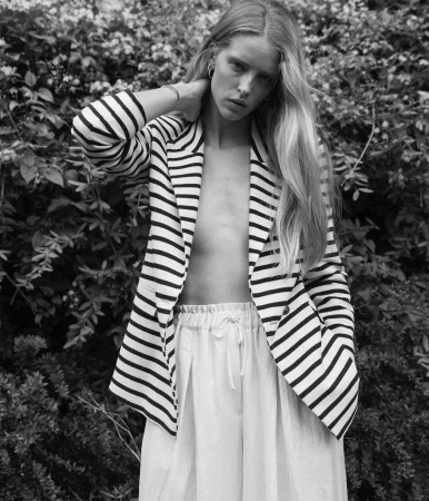 Black and white portrait of a woman with long blonde hair wearing a striped blazer and standing against a leafy background.