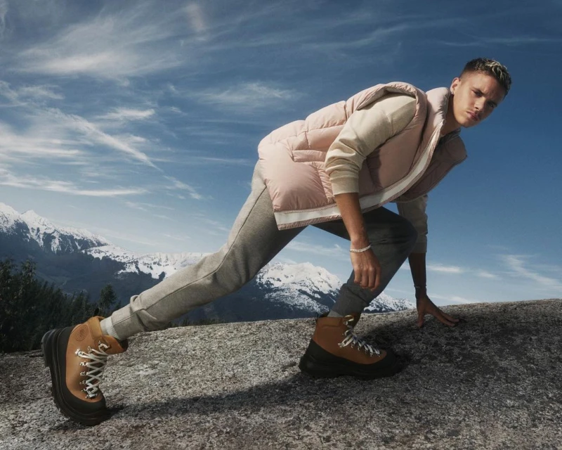 Male model poses outdoors wearing a vest, sweatshirt, sweatpants, and boots, with a mountain range and blue sky in the background.
