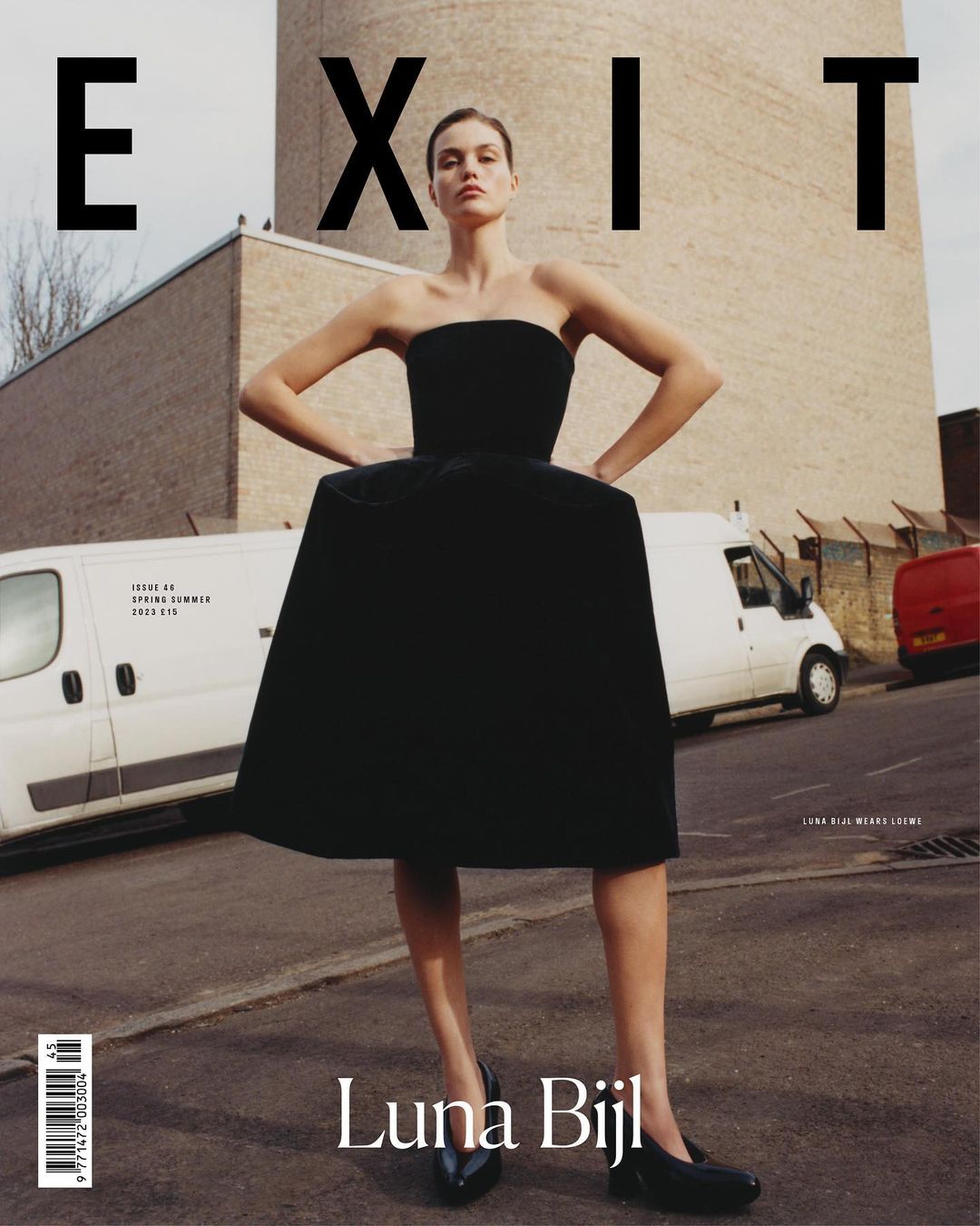 A woman in a black dress stands on a city street near parked vans, posing for a fashion shot. The image is used as a magazine cover.