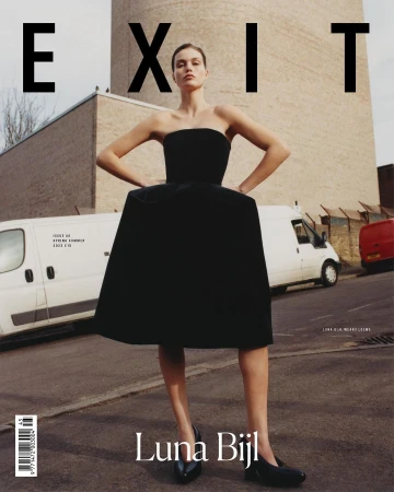 A woman in a black dress stands on a city street near parked vans, posing for a fashion shot. The image is used as a magazine cover.
