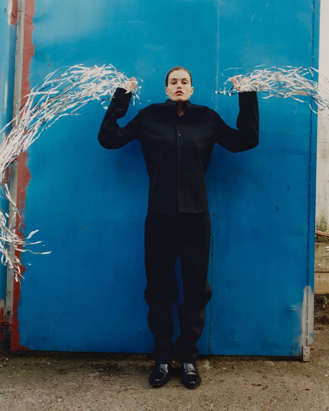 Fashion shot of a young adult woman in dark clothing standing in front of a blue wall, holding silver tinsel garlands overhead.