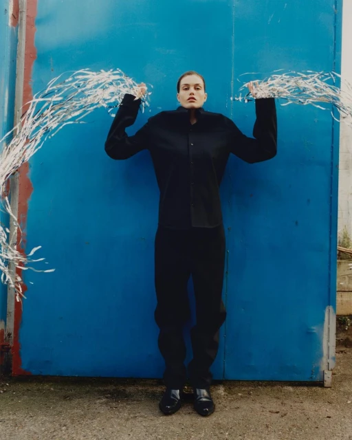 Fashion shot of a young adult woman in dark clothing standing in front of a blue wall, holding silver tinsel garlands overhead.