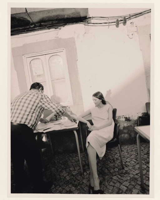 Black-and-white photo in an indoor setting: a woman seated while a man in a plaid shirt stands beside a table covered with papers.