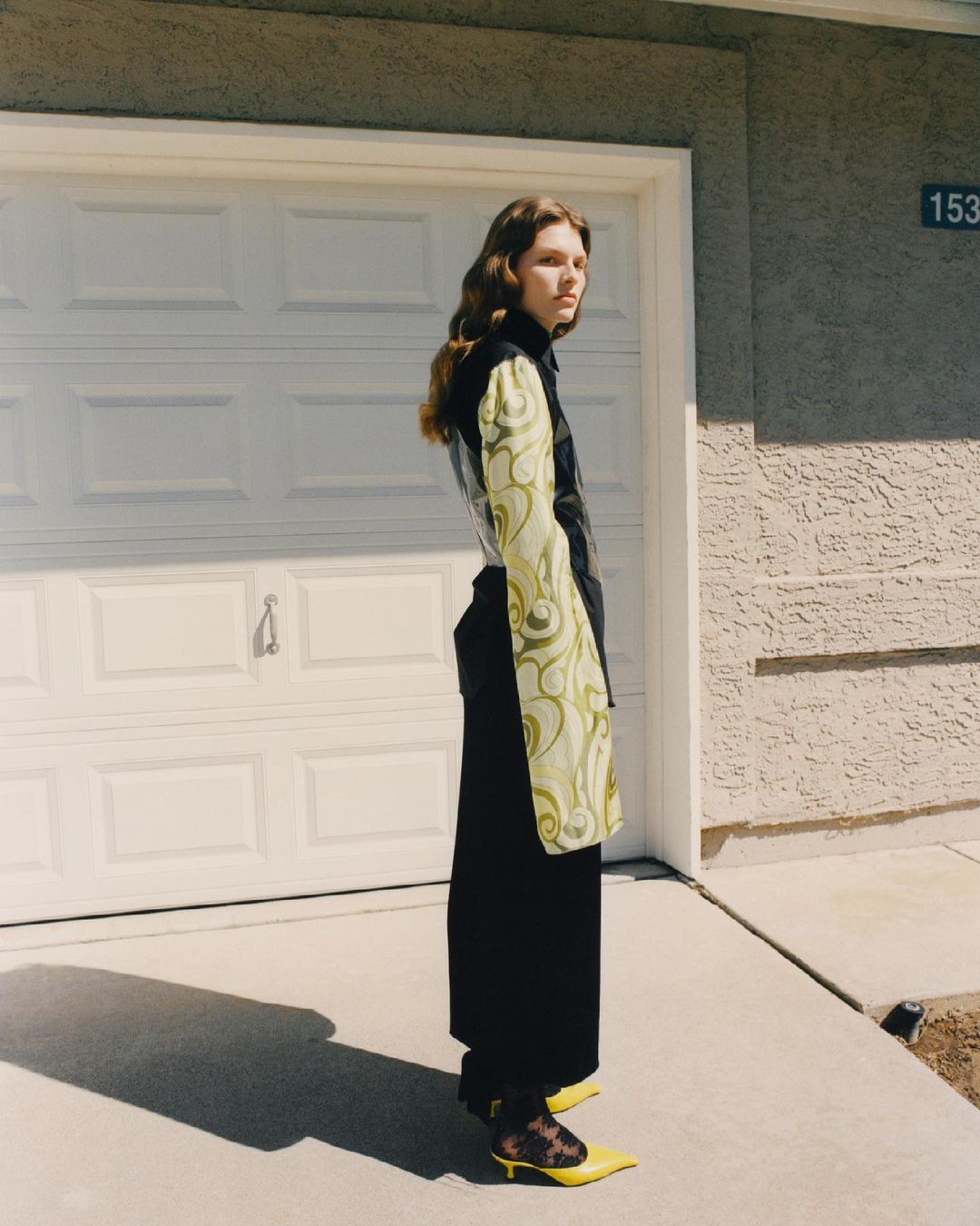 A woman with long brown hair is posing in front of a closed white garage door, wearing a black top, wide-leg pants, and yellow heels.