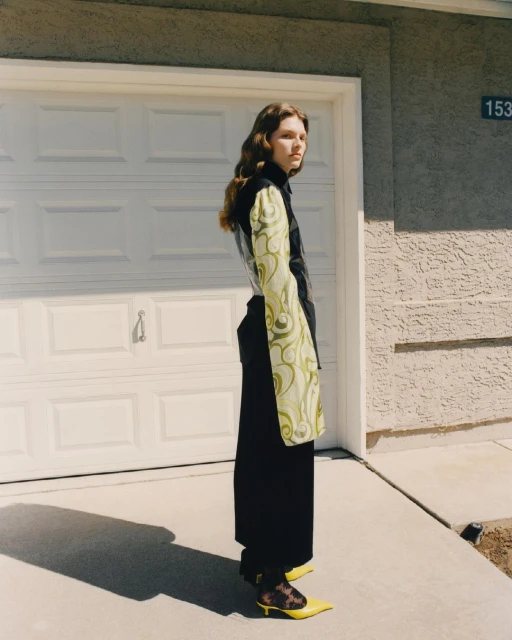 A woman with long brown hair is posing in front of a closed white garage door, wearing a black top, wide-leg pants, and yellow heels.