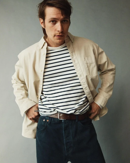 Portrait of a young adult male in a cream-colored jacket over a striped shirt, with blue jeans, posing in a neutral studio setting.