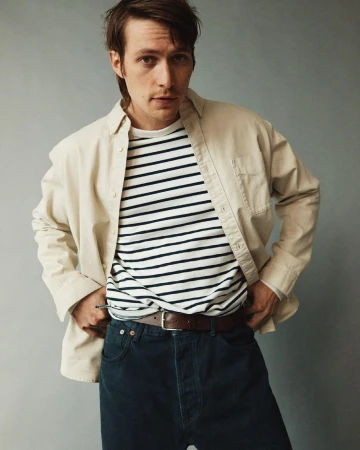 Portrait of a young adult male in a cream-colored jacket over a striped shirt, with blue jeans, posing in a neutral studio setting.
