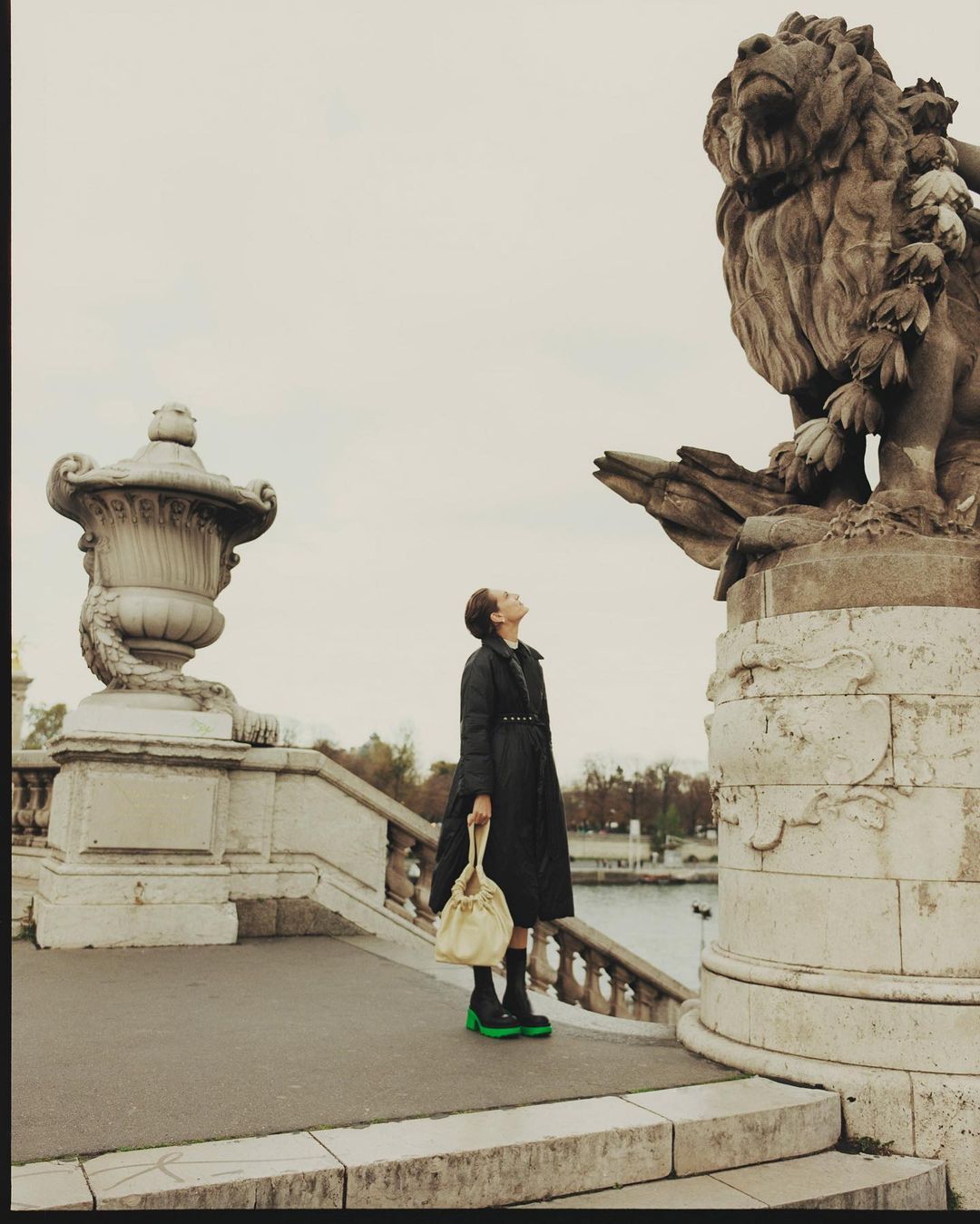Woman stands in front of lion sculpture in Paris, France, wearing long black coat, holding a light-yellow handbag, and gazing upward.