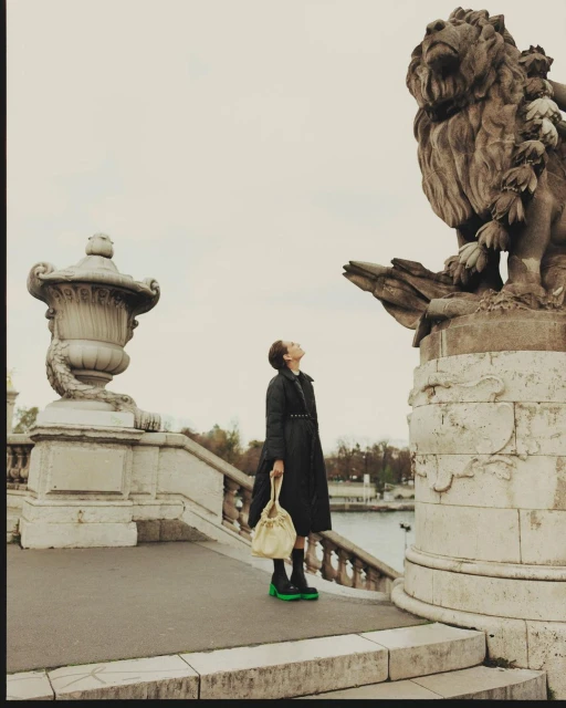Woman stands in front of lion sculpture in Paris, France, wearing long black coat, holding a light-yellow handbag, and gazing upward.