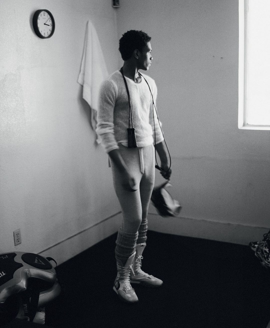Man in athletic wear stands in a room with a window and clock, holding boxing gloves and a jump rope in a black and white photograph.