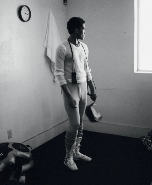 Man in athletic wear stands in a room with a window and clock, holding boxing gloves and a jump rope in a black and white photograph.