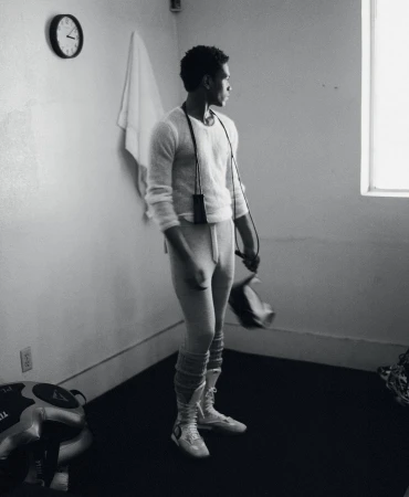 Man in athletic wear stands in a room with a window and clock, holding boxing gloves and a jump rope in a black and white photograph.