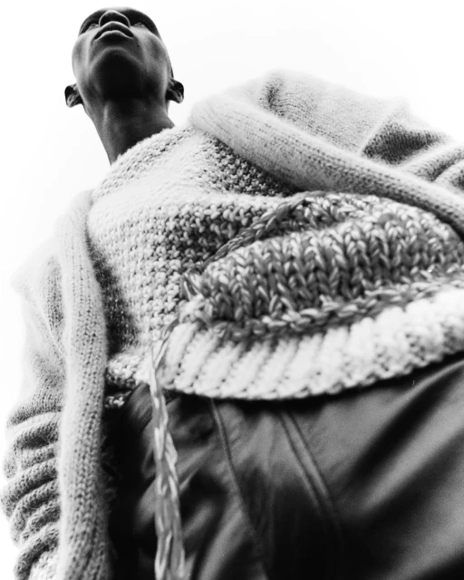 Monochrome low-angle shot of an African male wearing a white knitted sweater and black leather pants against a bright sky.