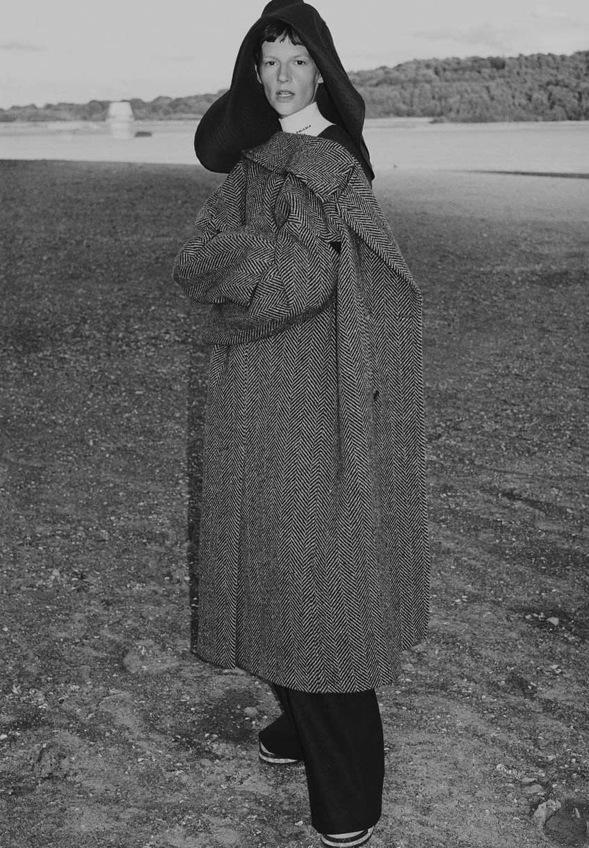 A person in an oversized coat with a hood poses in a field with trees in the background, fashion photography in black and white.