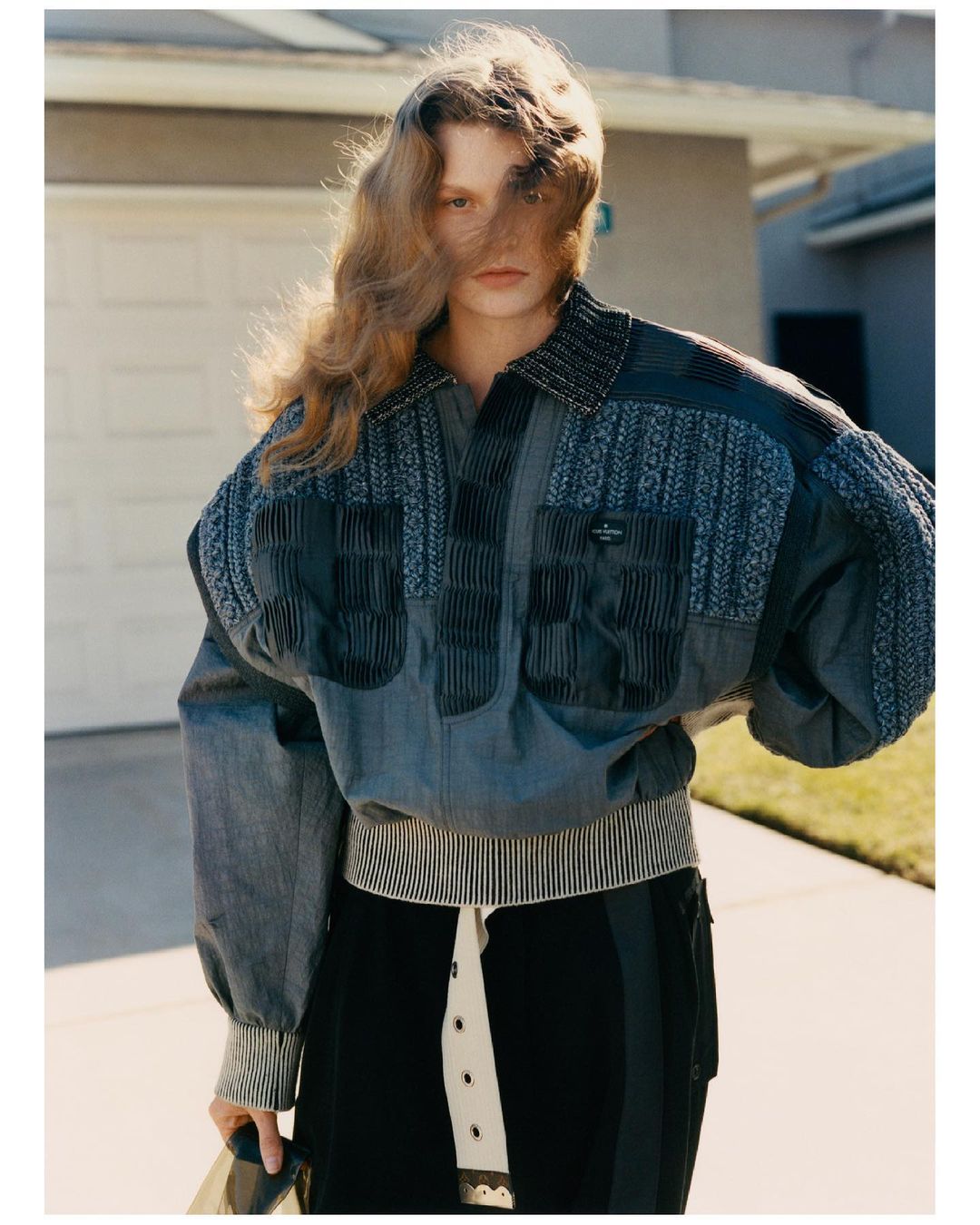 Portrait of a young adult woman wearing a patterned jacket, standing outdoors on a sidewalk in front of a suburban house on a sunny day.