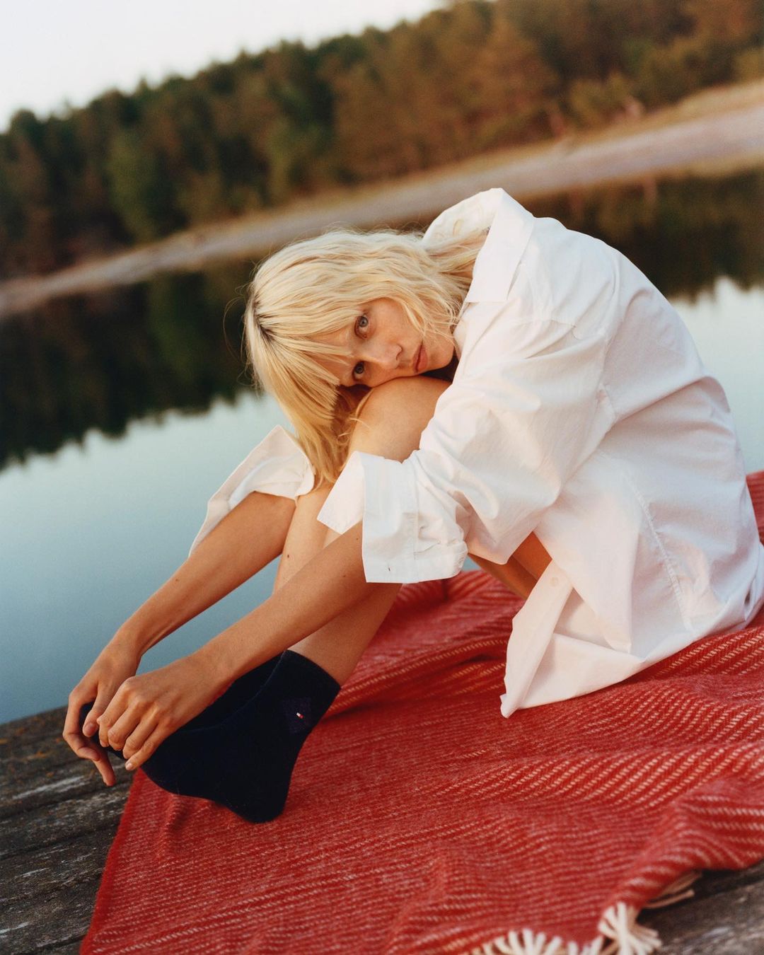 Blonde woman wearing a white button-down shirt sitting on a red textured blanket outdoors near water by Misha Taylor