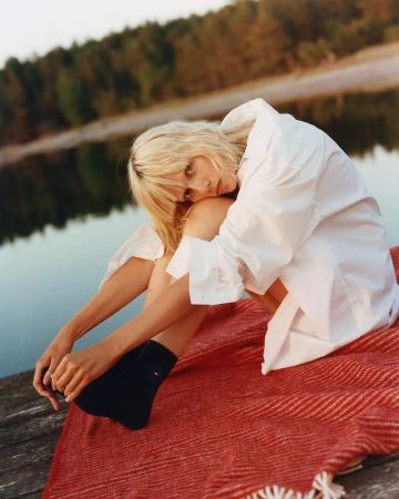 Blonde woman wearing a white button-down shirt sitting on a red textured blanket outdoors near water by Misha Taylor
