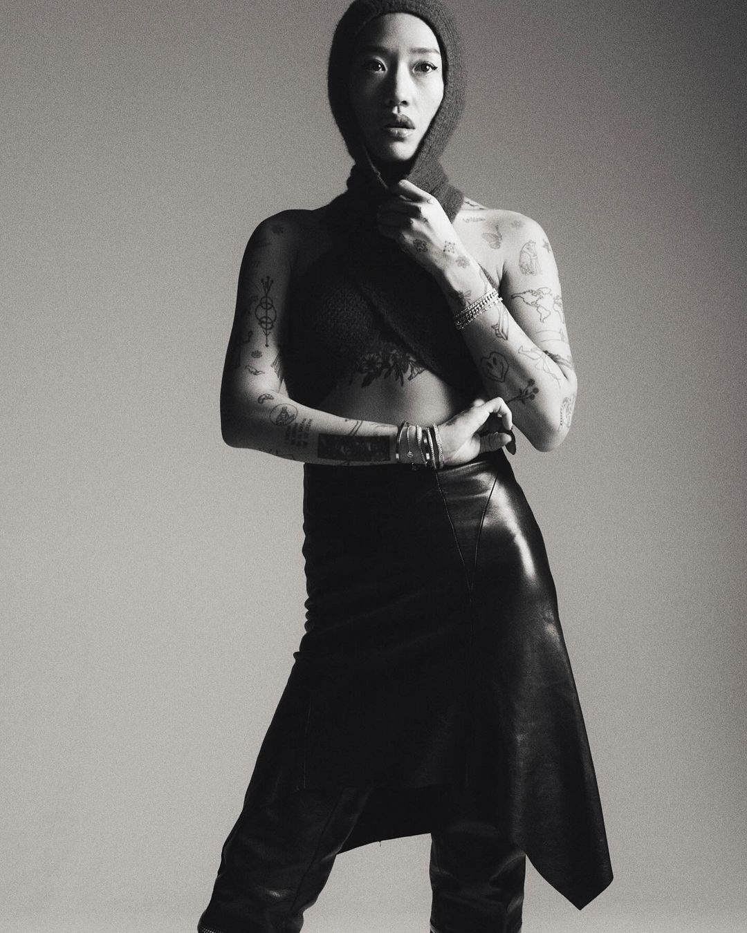 Monochrome portrait of a tattooed woman in a knit hood and leather skirt, posing against a plain gray background in a studio setting.
