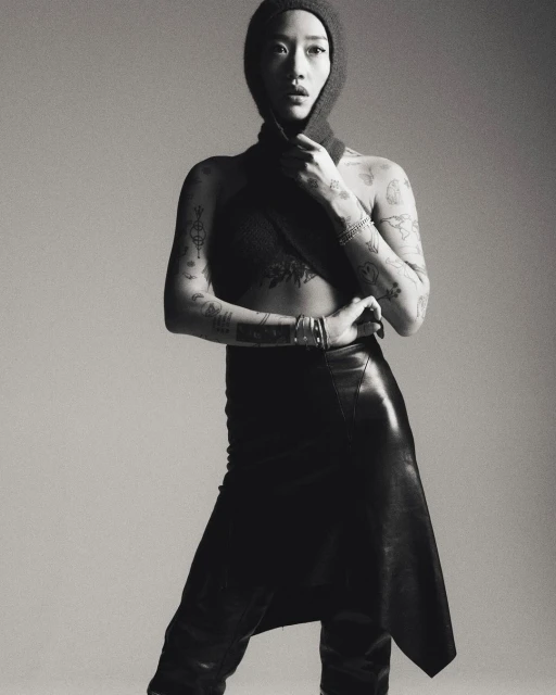 Monochrome portrait of a tattooed woman in a knit hood and leather skirt, posing against a plain gray background in a studio setting.