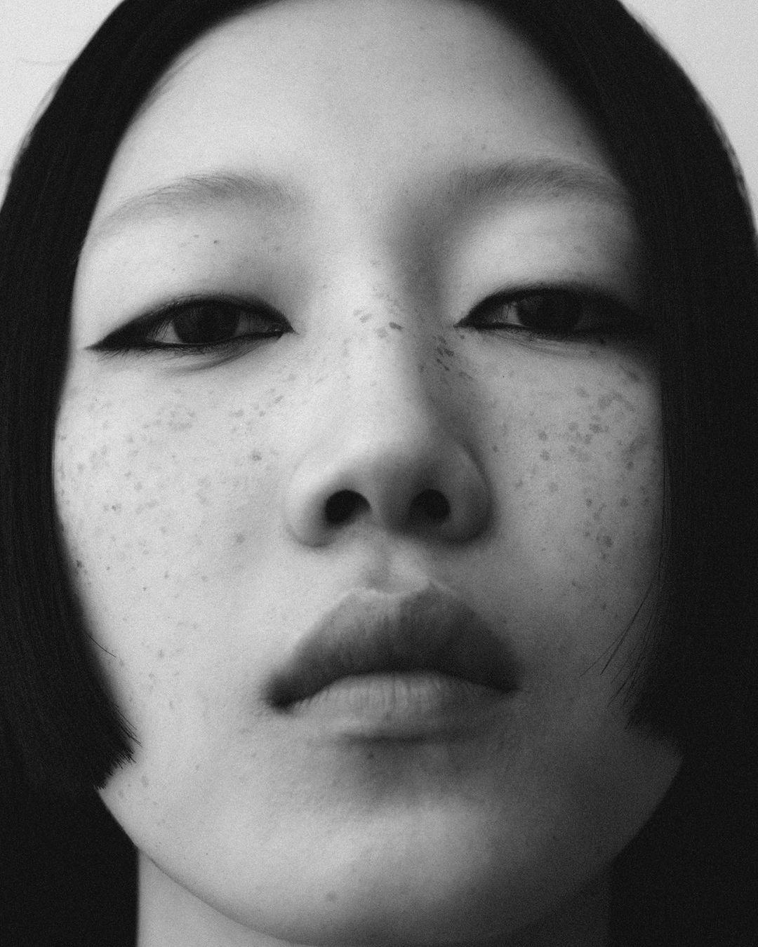 Striking black and white close-up of a woman's face showcasing her freckled skin, dark eyeliner, and bob haircut against a white backdrop.