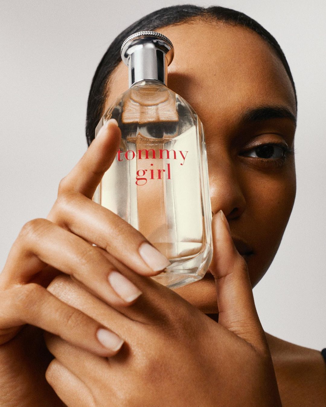 Close-up of a woman holding a bottle of Tommy Girl perfume, showcasing its clear glass and pink lettering against a neutral backdrop.