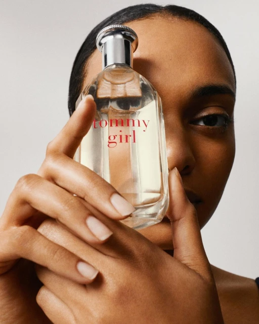 Close-up of a woman holding a bottle of Tommy Girl perfume, showcasing its clear glass and pink lettering against a neutral backdrop.