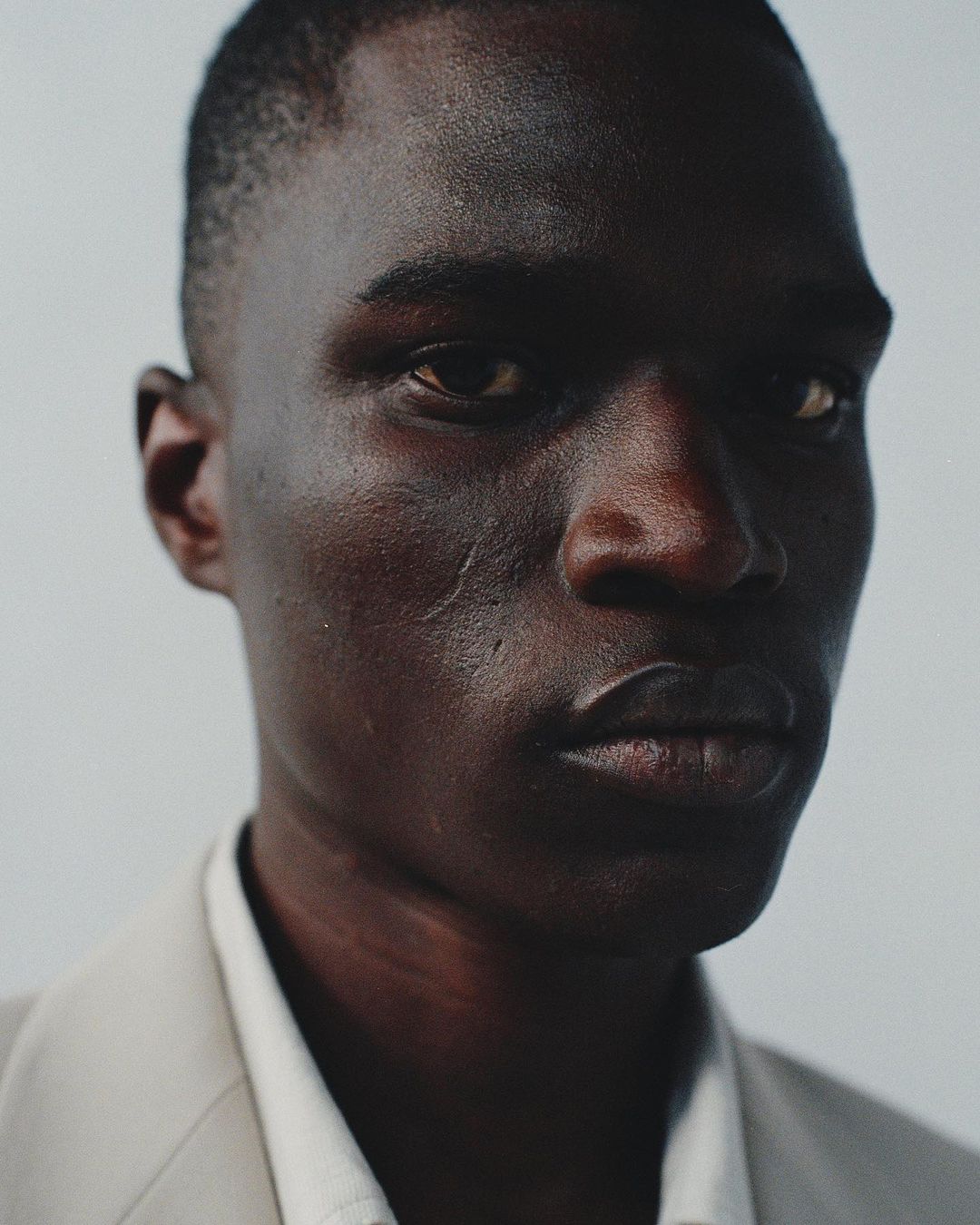 Close-up portrait of a Black man with short dark hair wearing a light suit jacket and white collared shirt against a plain background.
