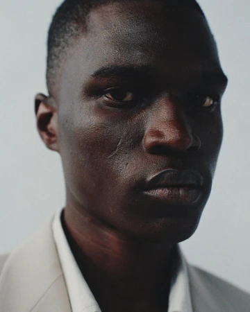 Close-up portrait of a Black man with short dark hair wearing a light suit jacket and white collared shirt against a plain background.