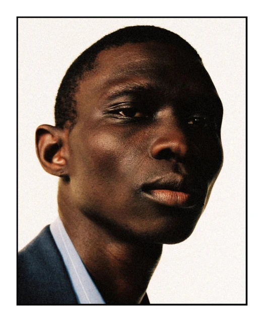 Close-up portrait of an adult Black male wearing a blue blazer, looking directly at the camera with a serious facial expression.